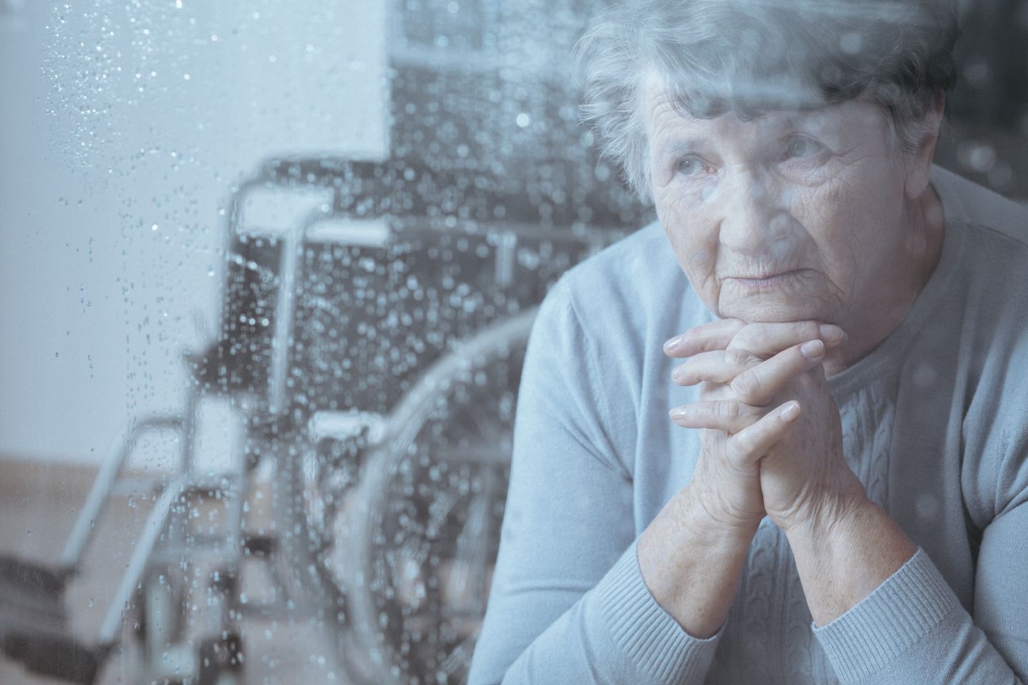 Elderly white woman with sad look on her face, hands folded with chin resting on them, looking out a window with a wheelchair in the background
